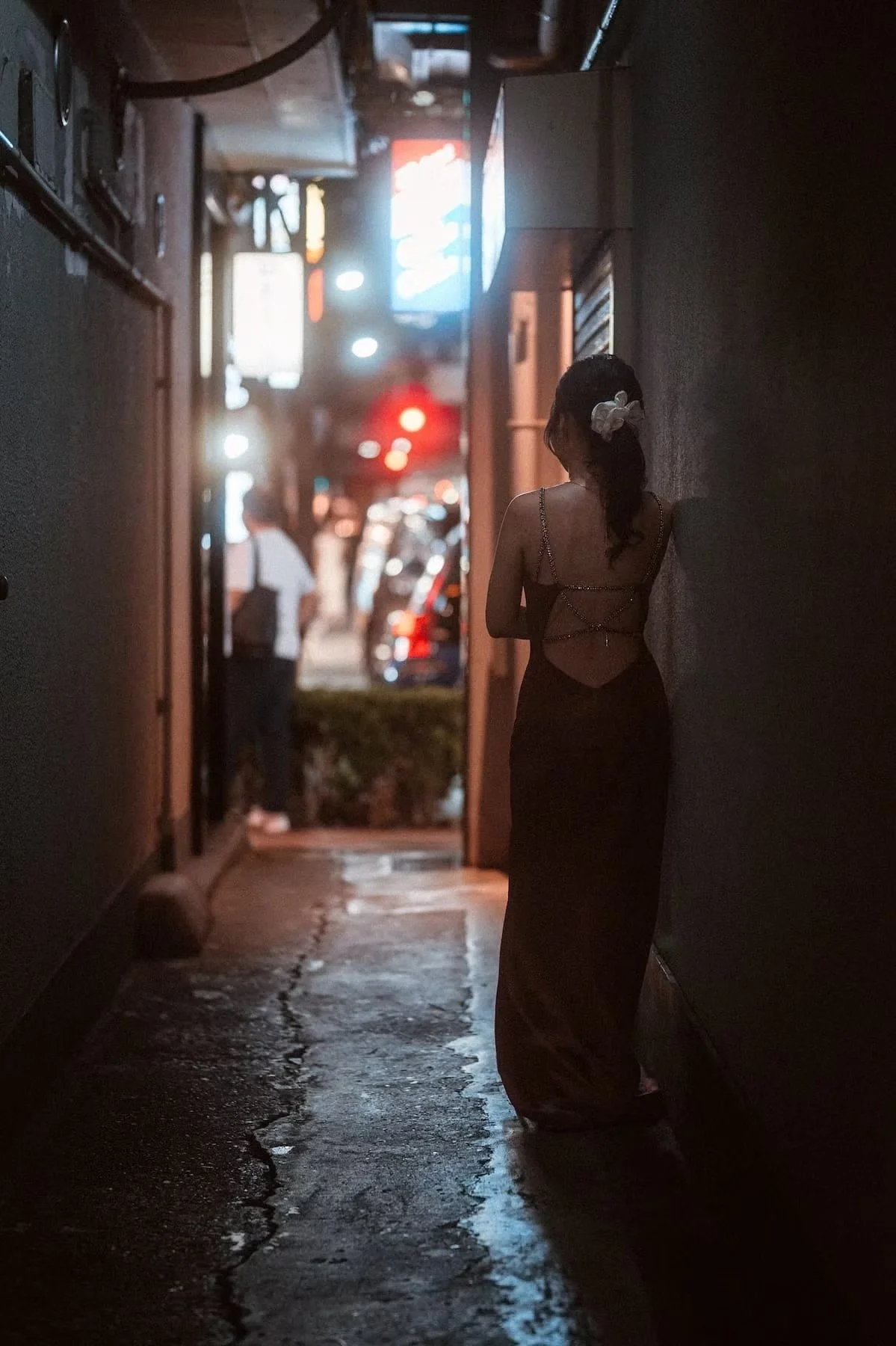 A hostess woman in Ginza Tokyo with a white bow in her hair, wearing a dark dress with a strap back, leaning against a wall in a dimly lit alleyway at night.