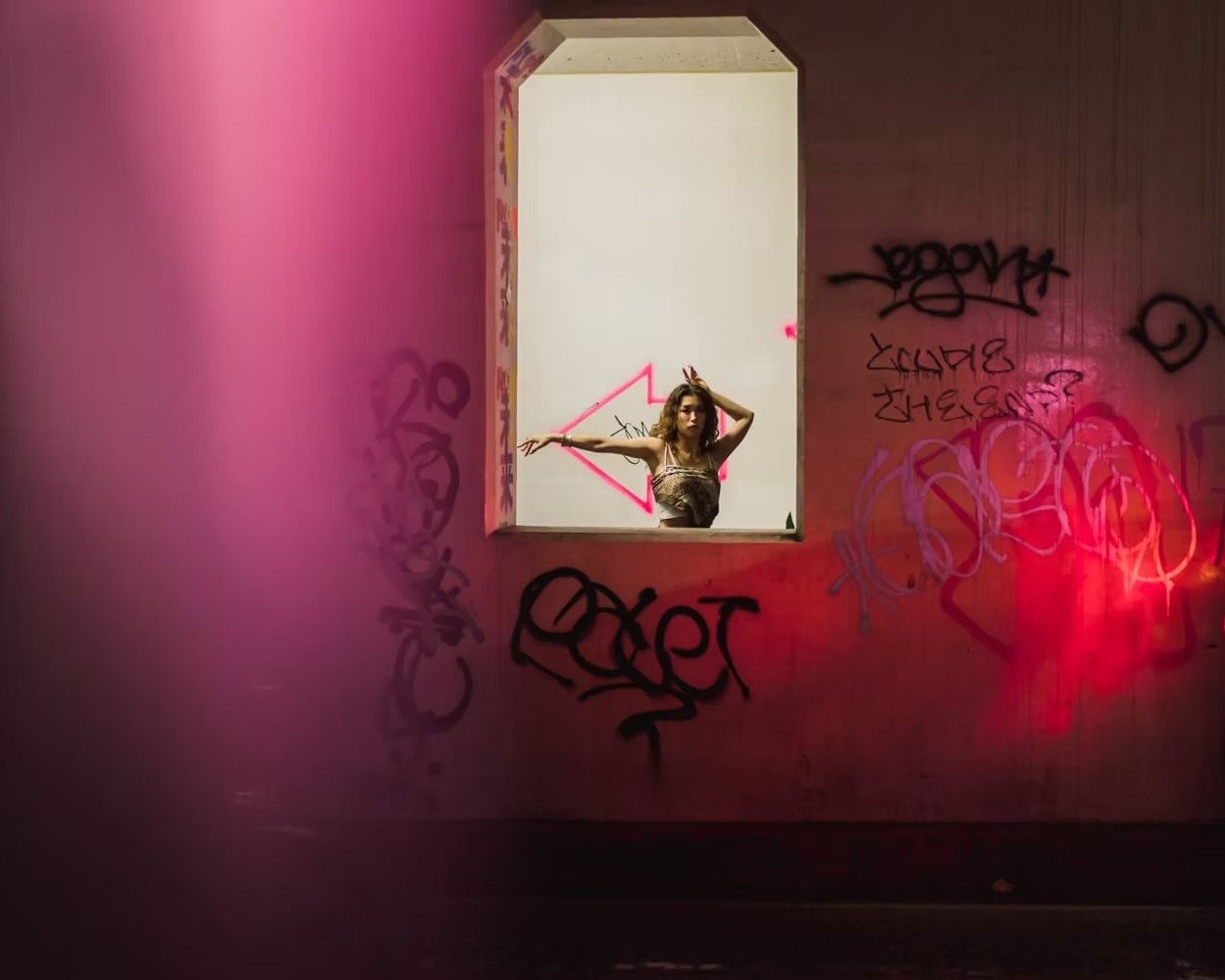 A woman with wavy hair wearing a dark top, posing at a window with graffiti-covered pink walls around it. The window has a pink neon arrow painted on the inside wall visible behind her shot in Shibuya.