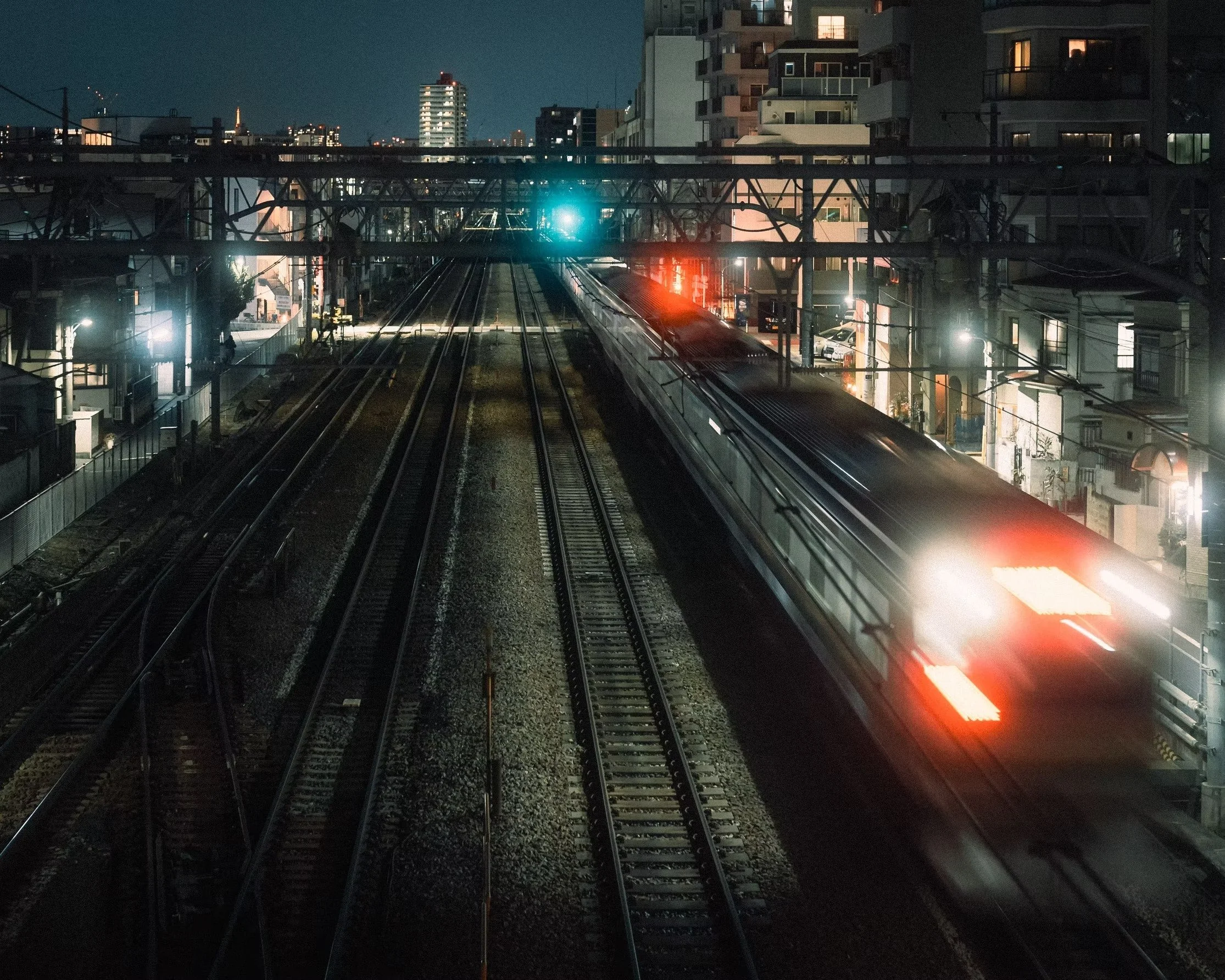 Nighttime view of train tracks from Uguisudani station in Tokyo with a train on the right side, illuminated buildings in the background, and city lights reflecting off the tracks.