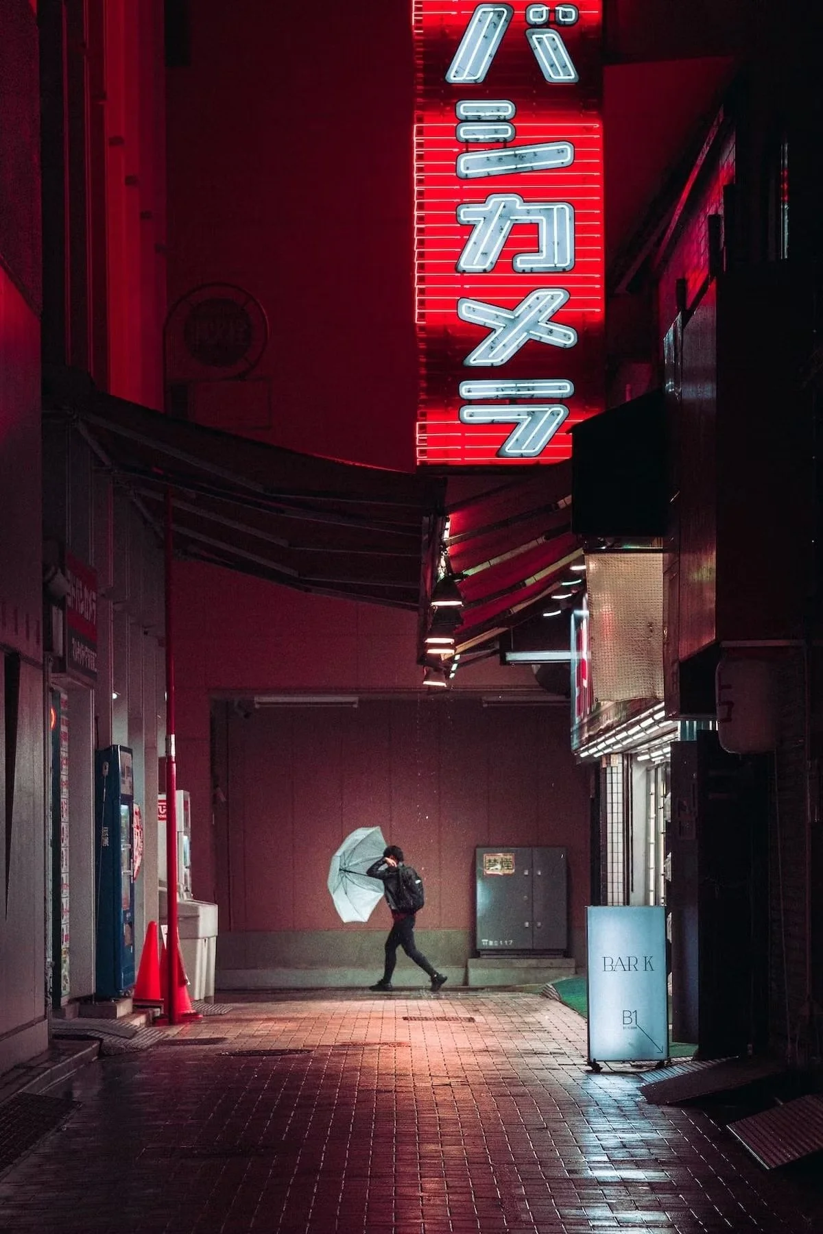 A person walking with an umbrella at night under a neon red and white sign with Japanese kanji characters, near a street corner with various signs and a vending machine.