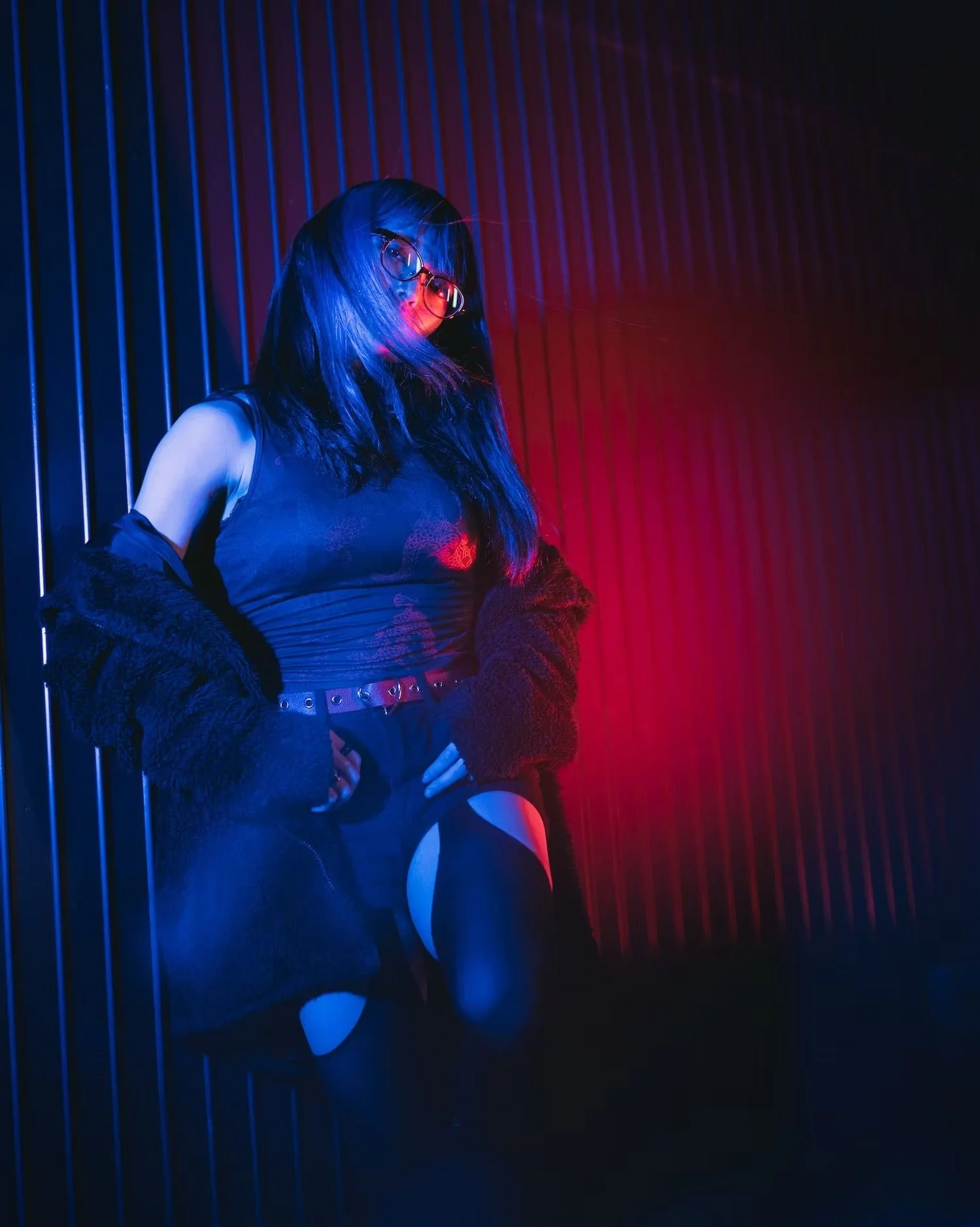 Girl with long hair, glasses, wearing a black outfit, illuminated by blue and red neon lights, standing in front of a striped wall.