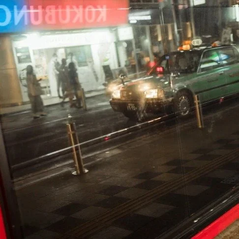 A city street at night is reflected in a large window, showing a green taxi with headlights on and pedestrians walking past a bright storefront.