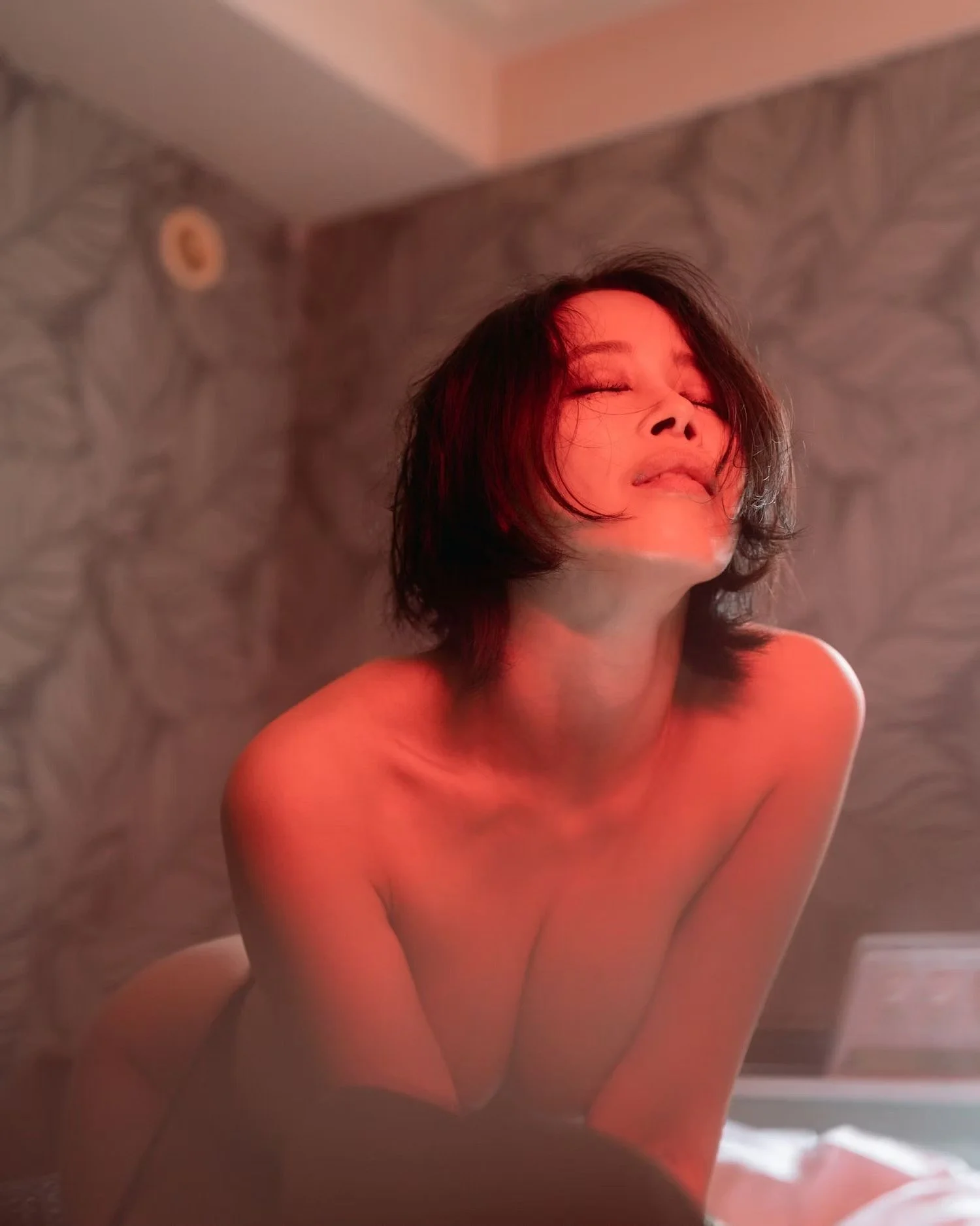 A woman with dark hair and closed eyes, smiling slightly, with her head tilted back and shoulders exposed, against a patterned wall with warm lighting.