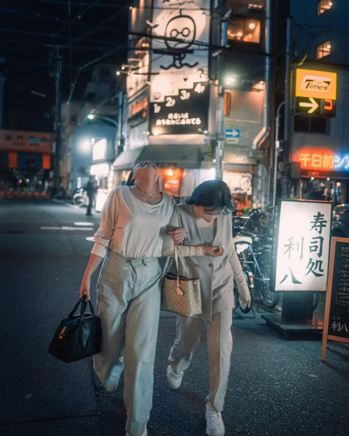 Two women walking arm in arm on a city street at night, illuminated by neon signs and streetlights in Osaka Japan. One woman carries a black handbag, the other holds a shopping bag.