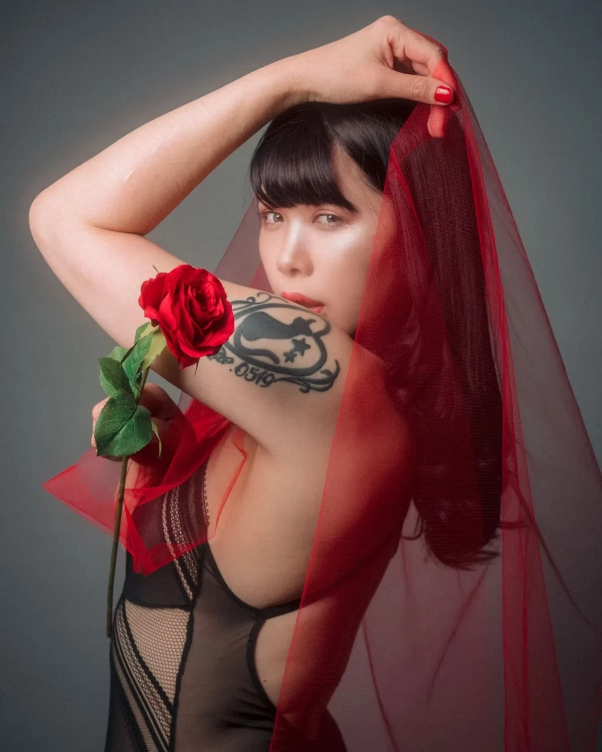 A model wearing a black lace outfit holds a red rose while draped in a red veil. The contrast between the red veil and the subtle blue background creates a striking image, adding a mysterious and sensual atmosphere to the scene.