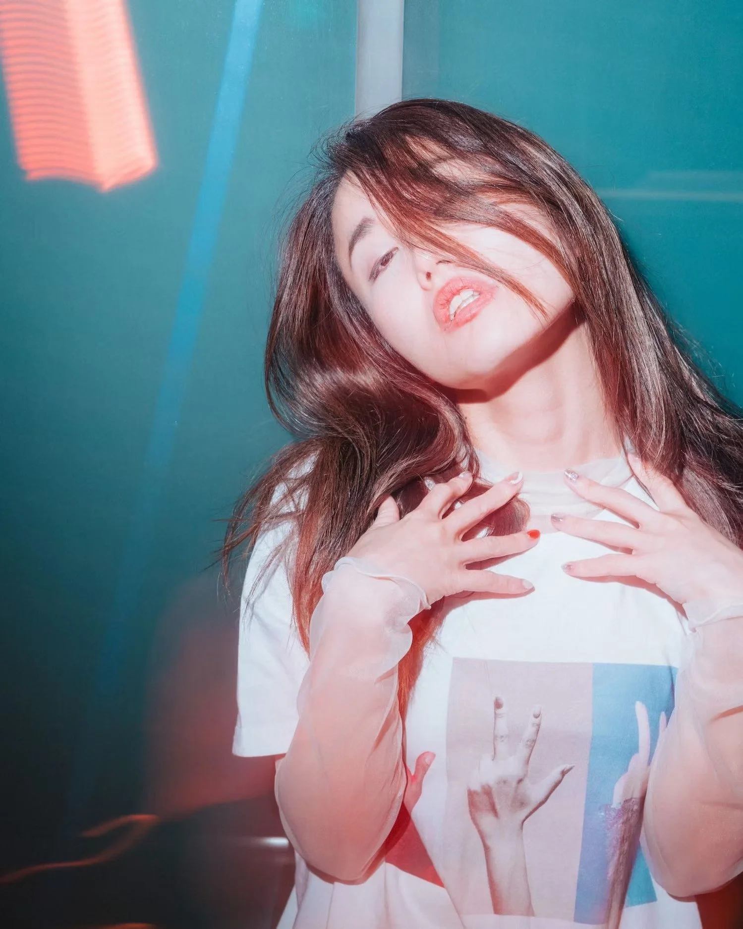 A woman with long brown hair styled with waves, partially covering her face, wearing a white top, holding her hands near her chest, with a vibrant, colorful background with pink, blue, and teal hues.
