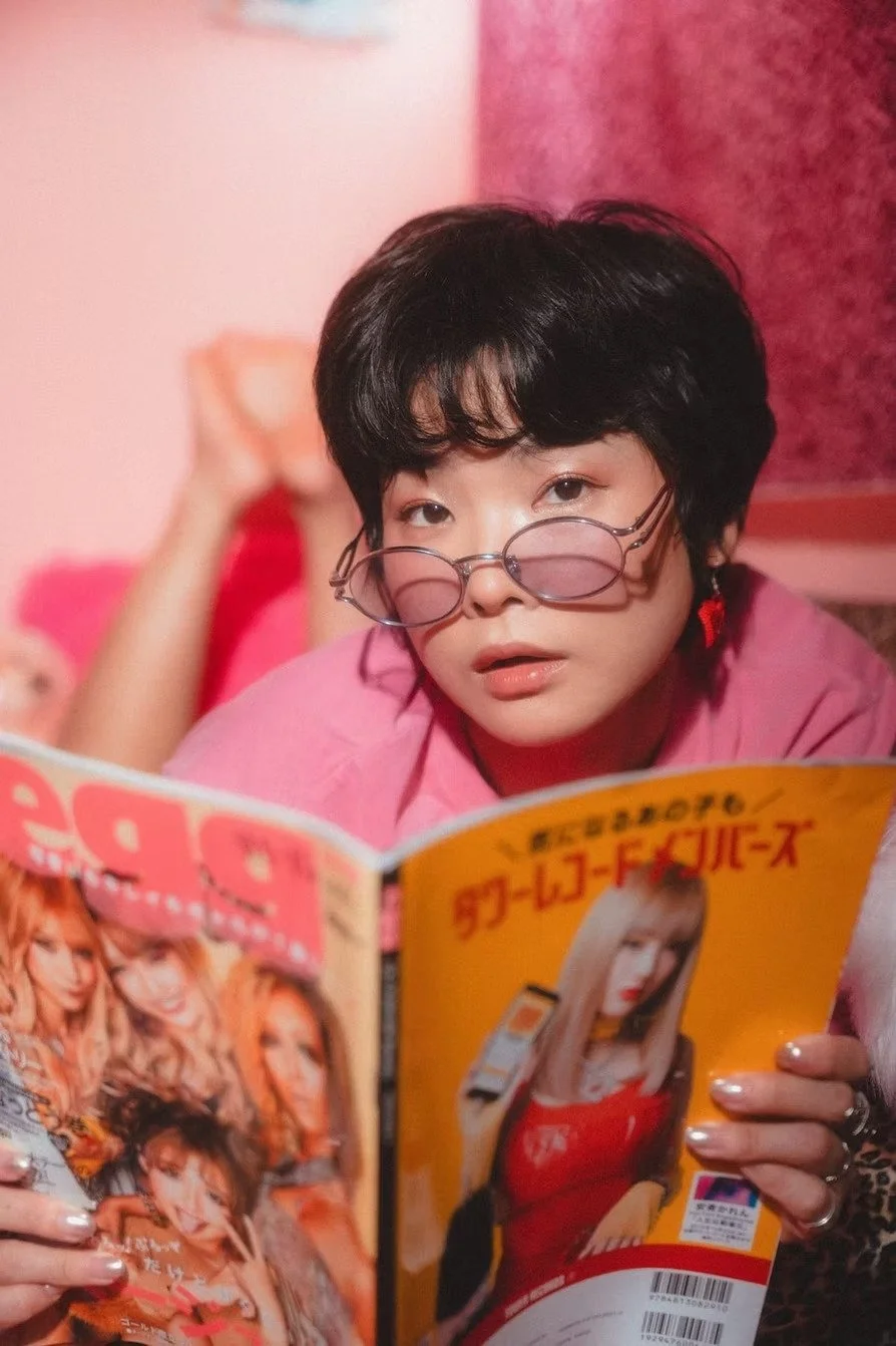 A person with short black hair wearing pink clothing and round glasses with tinted lenses, lying down and reading a fashion magazine with a yellow cover that features a woman with long blonde hair.
