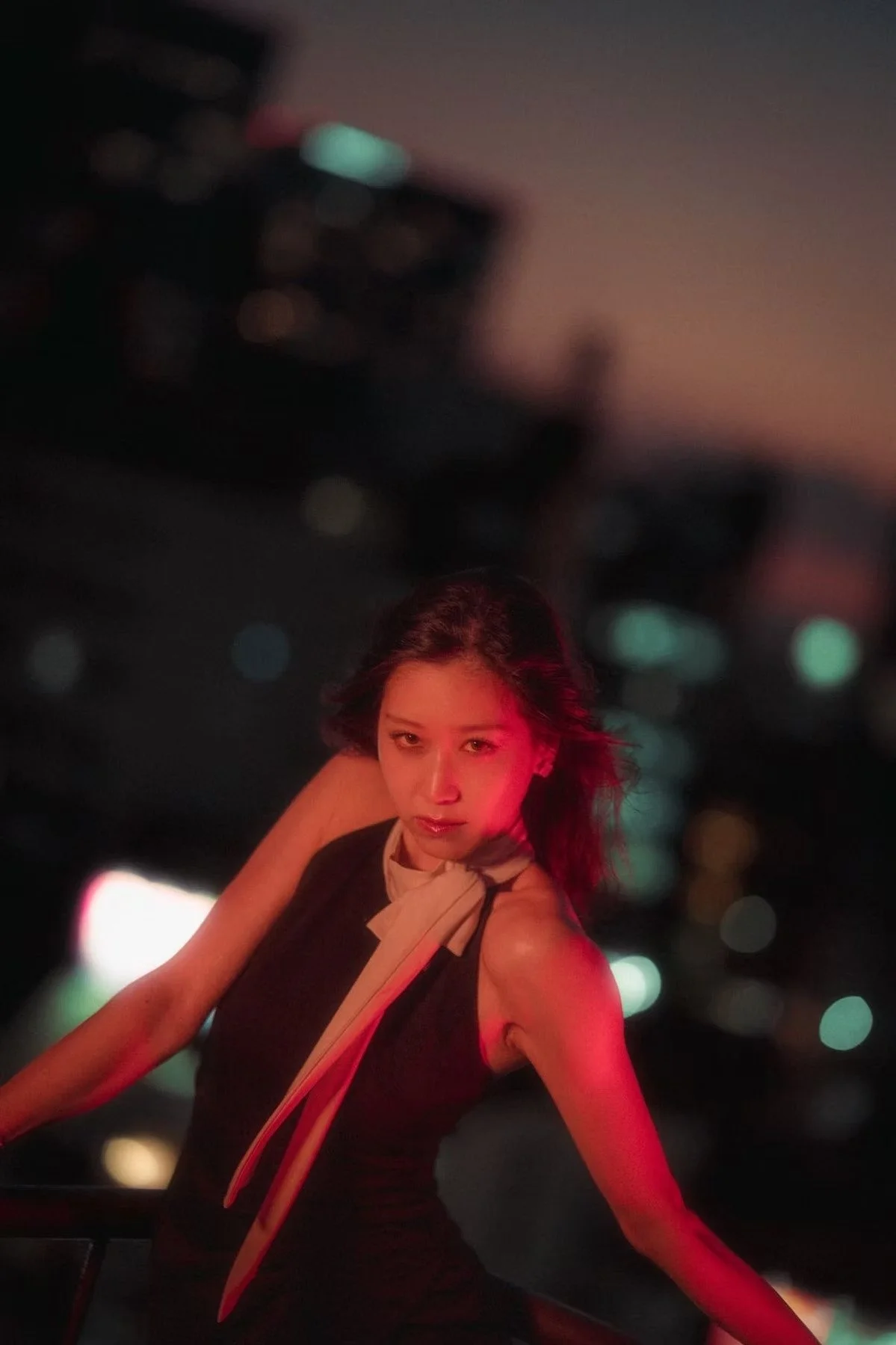 A young woman with shoulder-length hair poses at night with city lights in the background, illuminated by red lighting on a Tokyo rooftop.