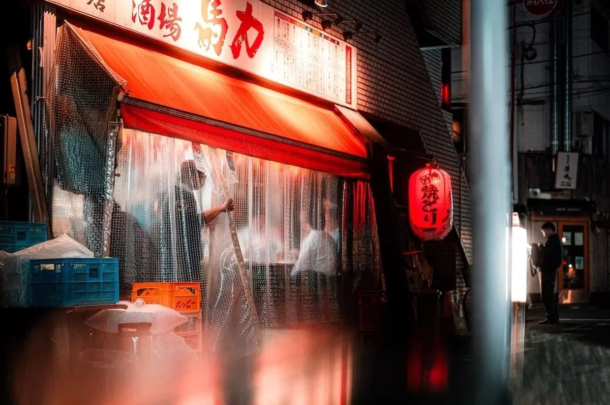 A Japanese street scene at night with a food stand or restaurant decorated with red lanterns and illuminated signage, with people inside and outside in Shimbashi Tokyo.