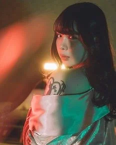A woman with black hair and bangs, wearing an off-shoulder top, looking over her shoulder with colorful lighting.