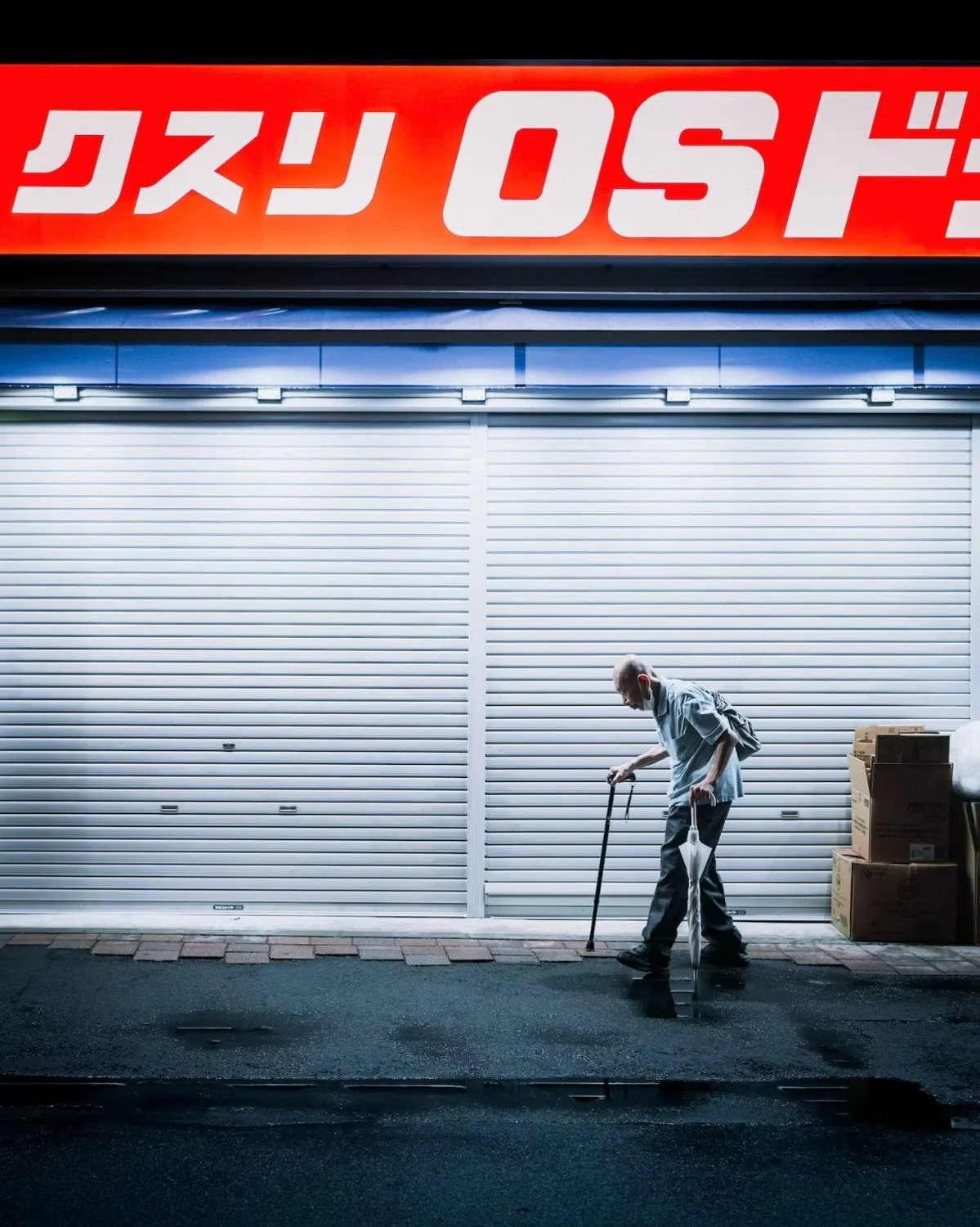 An elderly Japanese man with a cane walking past a closed storefront with a metallic shutter, stacks of cardboard boxes are on the right side of the scene in Tokyo. 