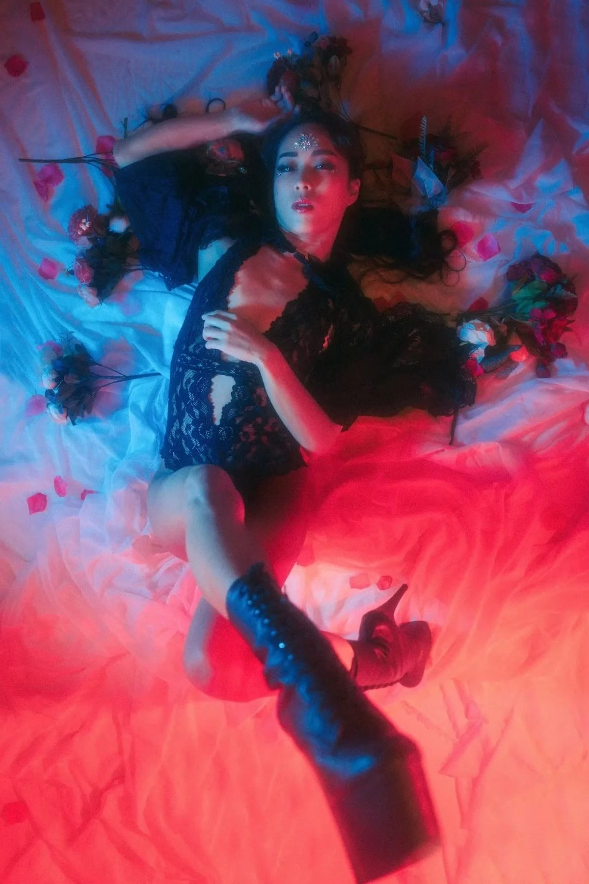 A model lies gracefully in a red and blue-lit environment, surrounded by mist. The vibrant lighting and soft movement create a sensual and dreamlike atmosphere, drawing attention to the model's posture and the dramatic lighting effects.