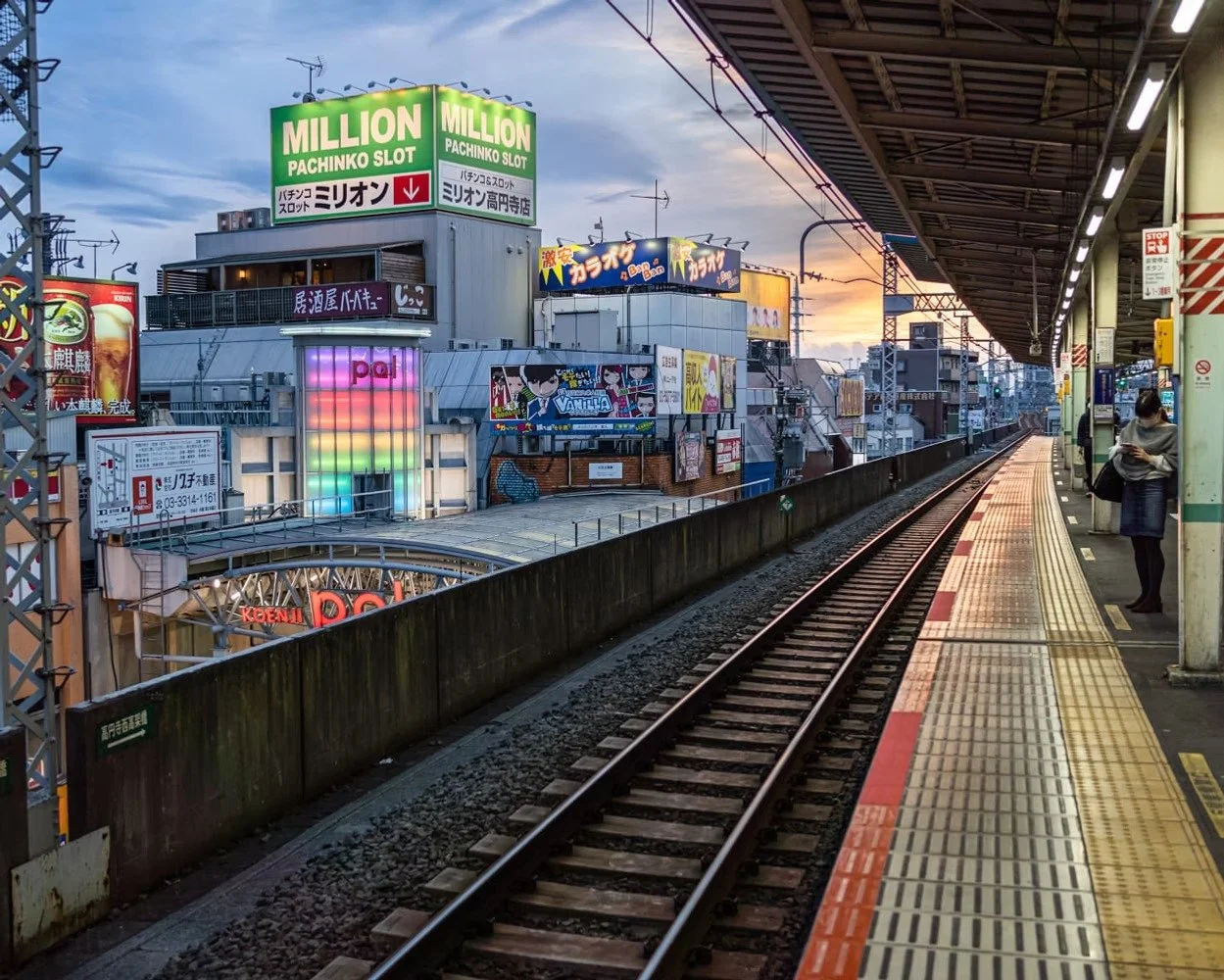 A city train platform during sunset with a woman waiting on the right, multiple colorful signs and billboards on buildings across the street, and the train tracks extending into the distance.