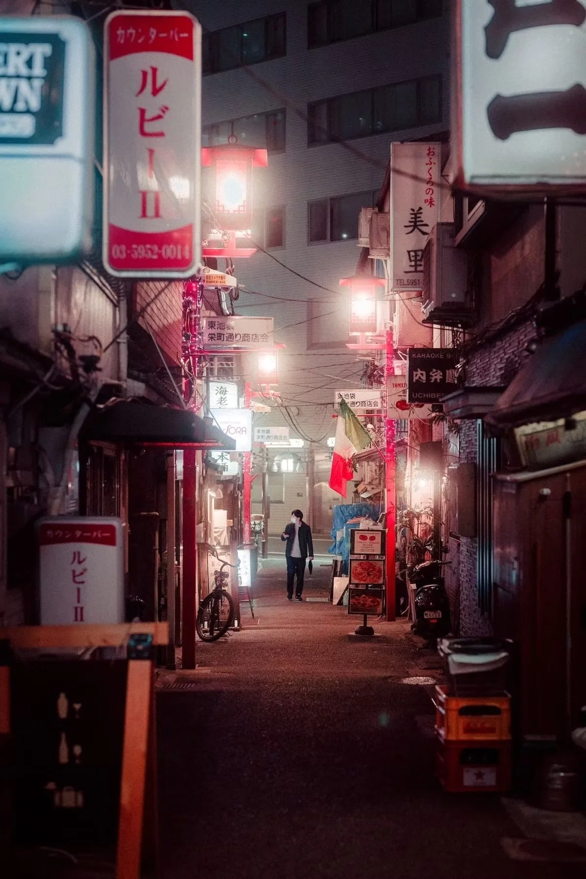 A narrow street at night illuminated by pink and white neon signs, with a man walking and wearing a mask, buildings on both sides, bicycles parked, and small shops with signs in Japanese. Ikebukuro.
