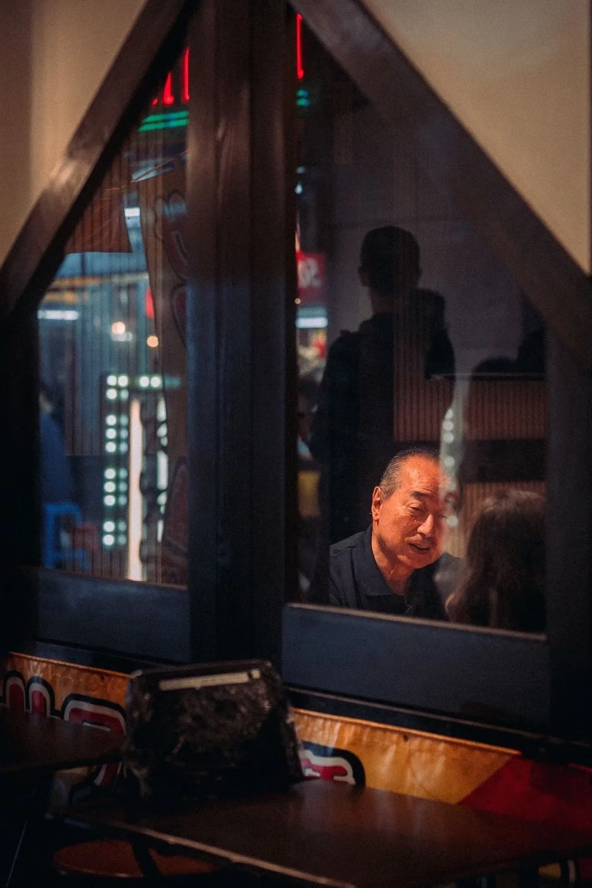 A reflection of a Japanese salaryman talking to a woman in a restaurant window at nighttime, with neon lights and other people visible in the background.