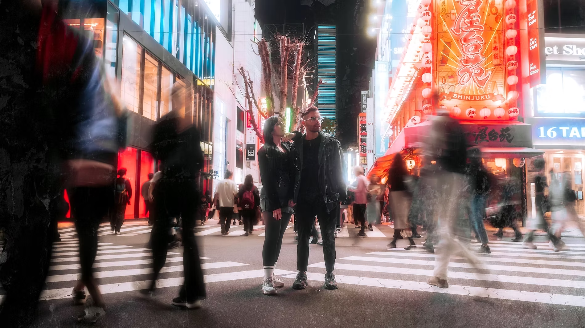 Night scene of busy pedestrian street in Tokyo, Japan, with neon signs and a large illuminated market sign reading 'Shinjuku.' Two people stand in the crosswalk, surrounded by blurred motion of other pedestrians.