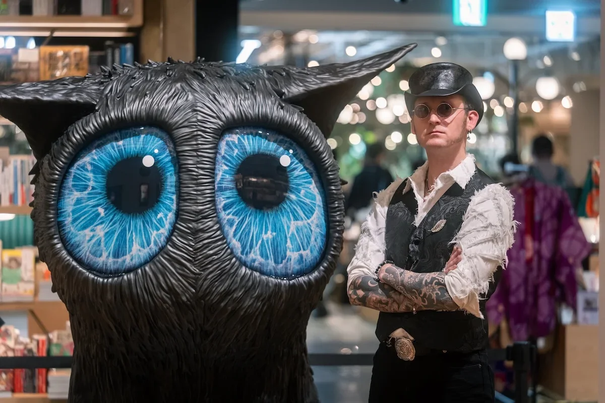 August Vilella
---
Creative environmental portrait of artist August Vilella standing behind one of his large black creature sculptures with striking electric-blue eyes. The photograph captures the artist's distinctive personal style alongside his ima