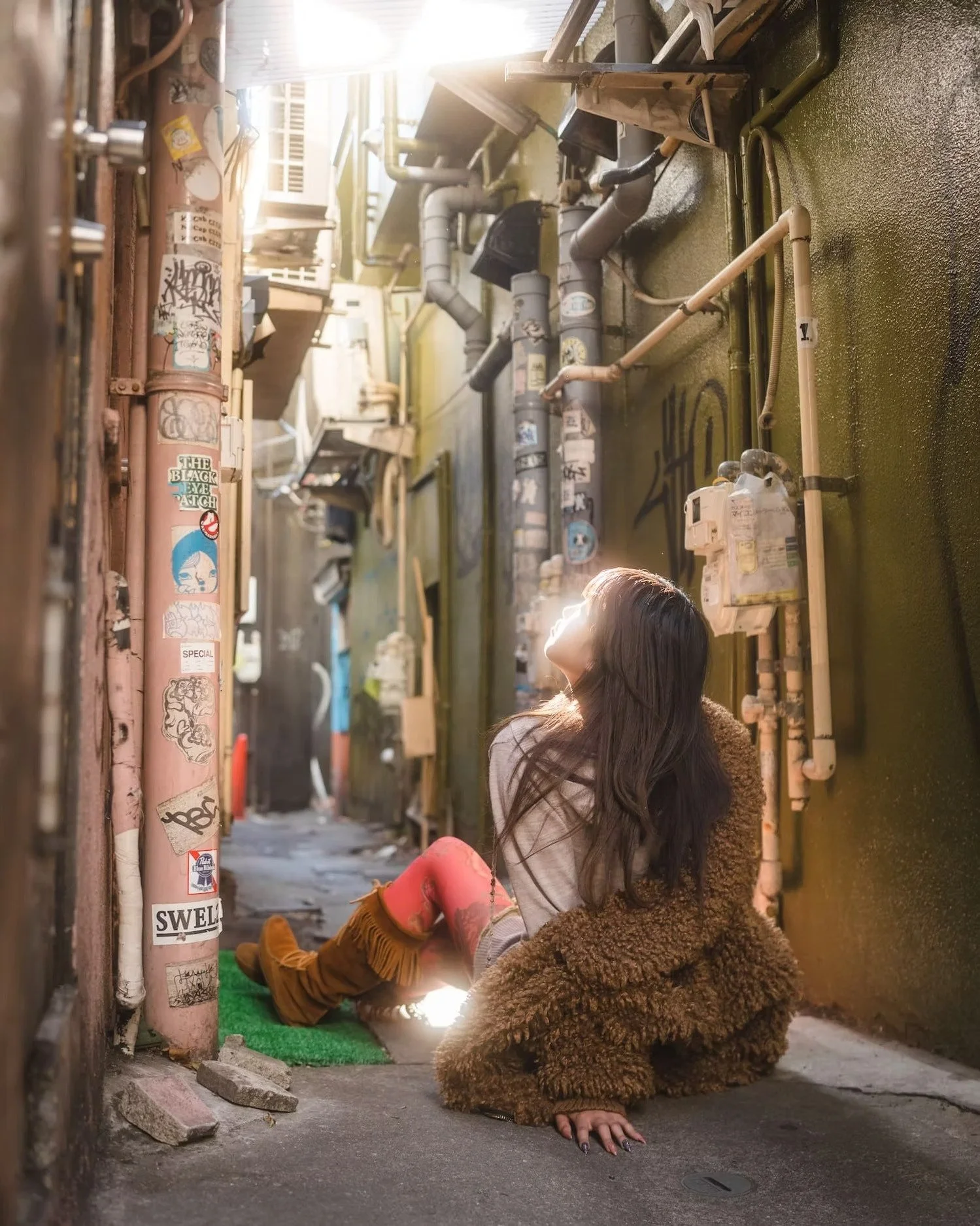 A woman with long dark hair sitting against a graffiti-covered wall in an alley, wearing a brown furry coat, tan boots, and red pants. She is looking towards the sunlight with her head tilted back, next to exposed pipes and graffiti-covered surfaces.