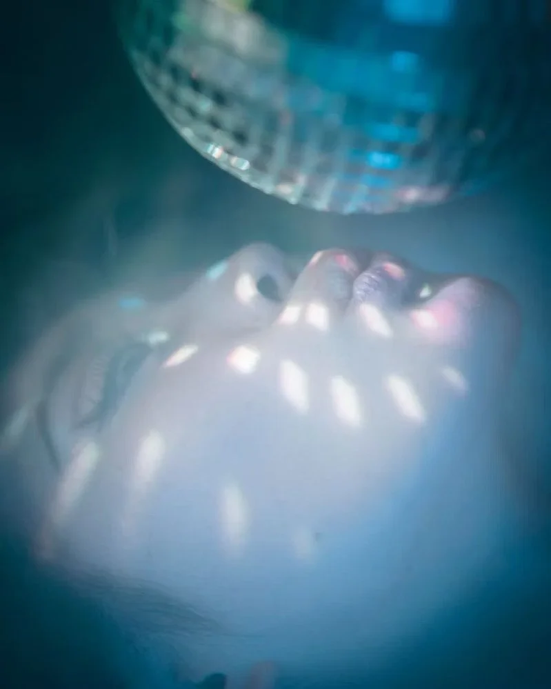 Close-up of a person's face illuminated by light reflecting off a disco ball, with a blue background.