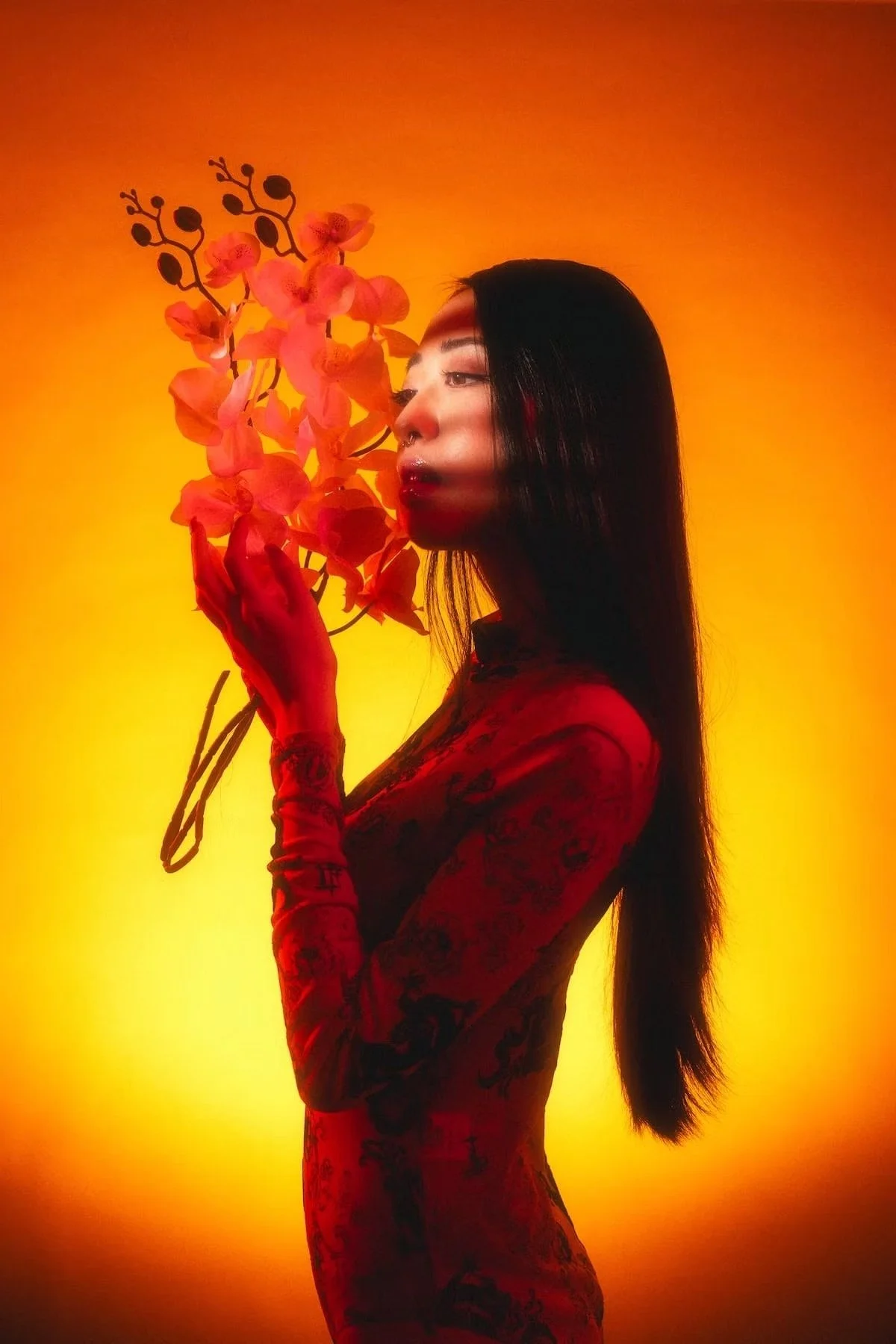 A woman with long black hair, wearing a red patterned dress, holding a branch with pink orchids, standing against a warm orange and yellow gradient background.
