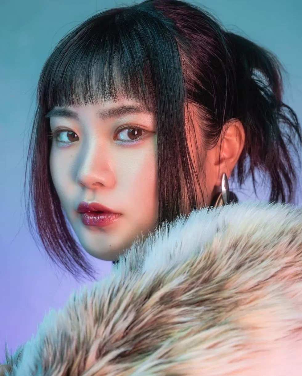 A model with a fur collar looks directly at the camera, with her hair styled in a way that emphasizes her striking facial features. The background is a mixture of warm reds and blues, giving the image a dramatic and bold vibe.