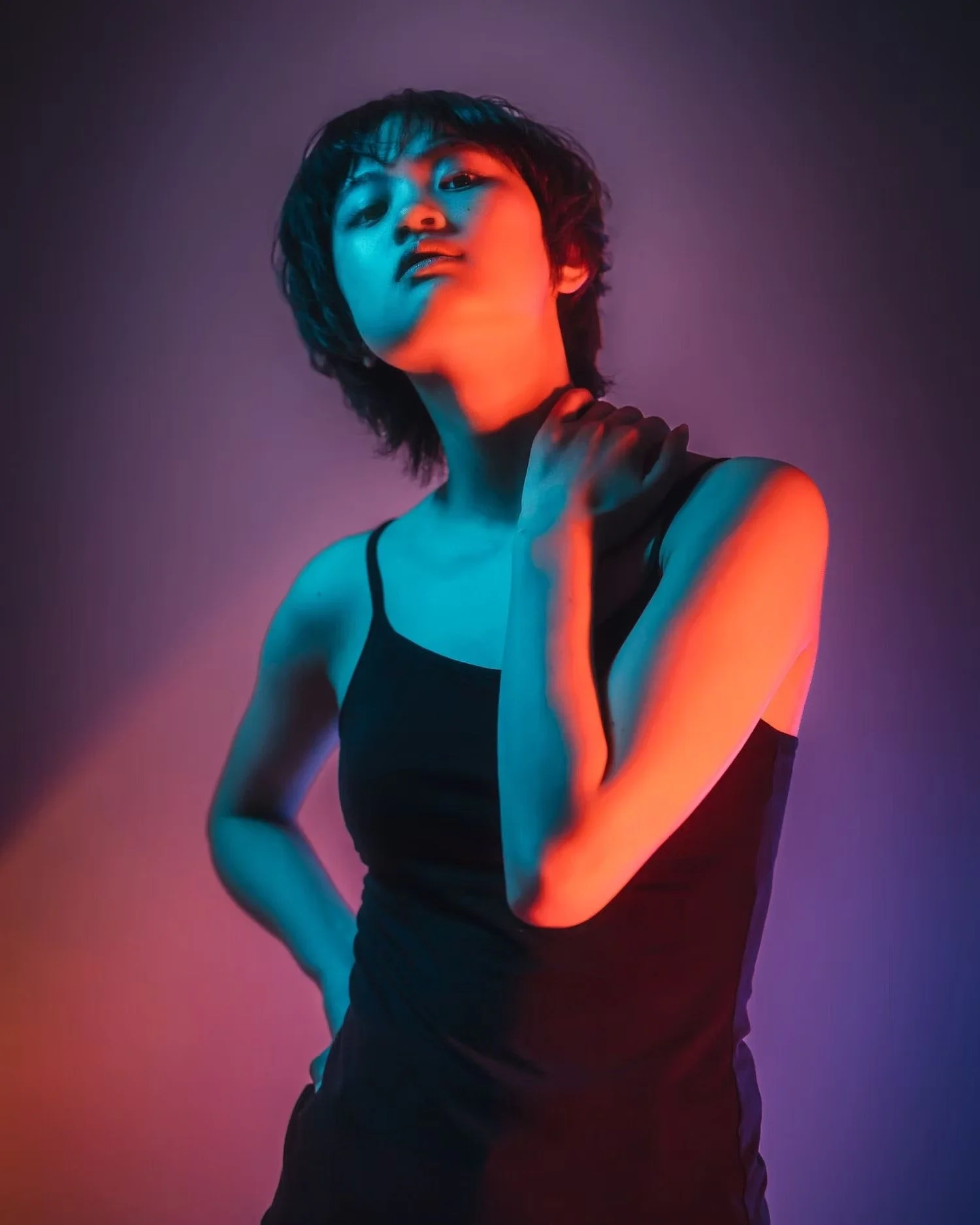 A young woman with short, wavy hair posing against a gradient background with pink and blue lighting. She is wearing a black sleeveless top and has her hand on her neck, looking at the camera with a confident expression.