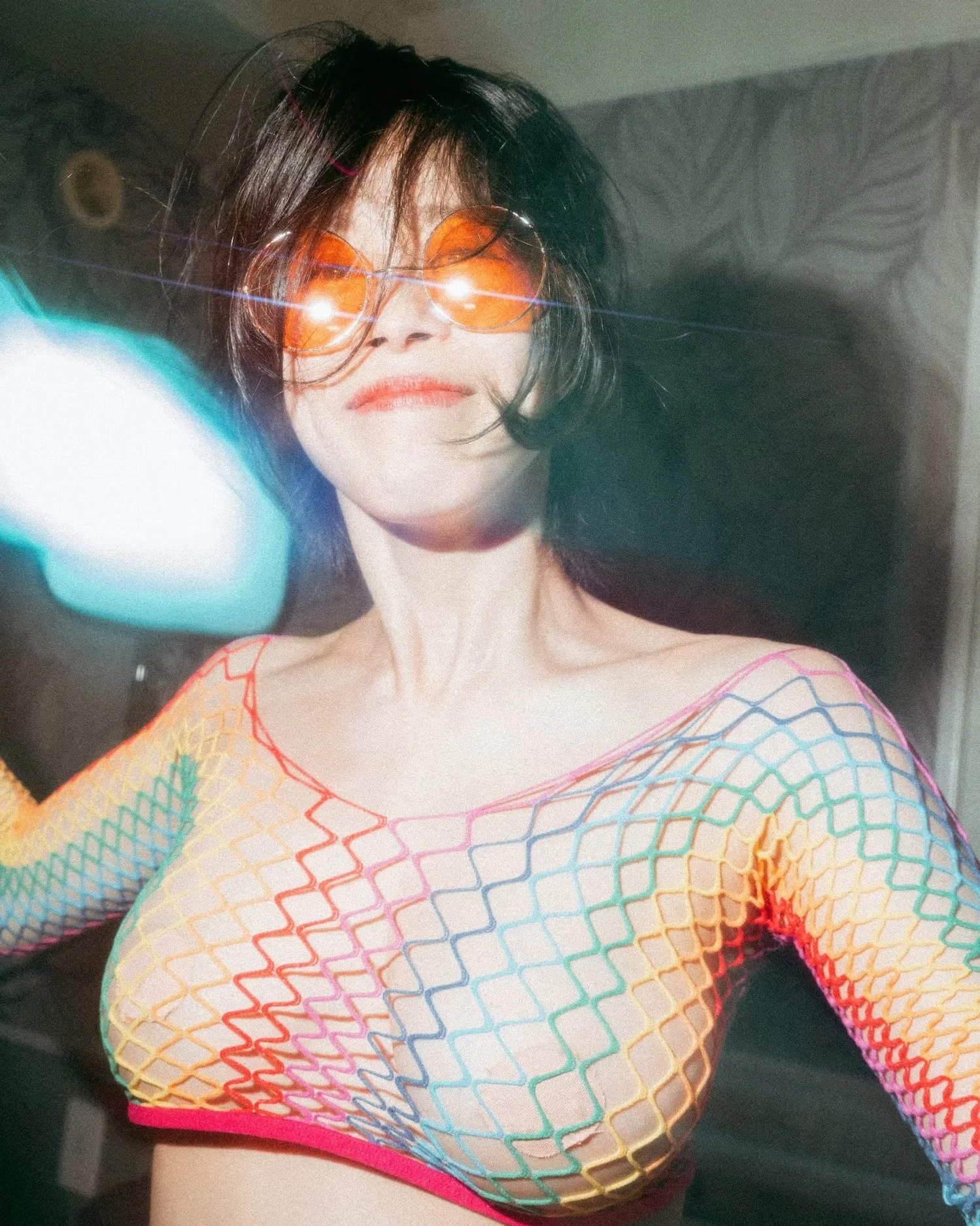 A woman wearing orange-tinted sunglasses and a colorful fishnet top with rainbow patterns.