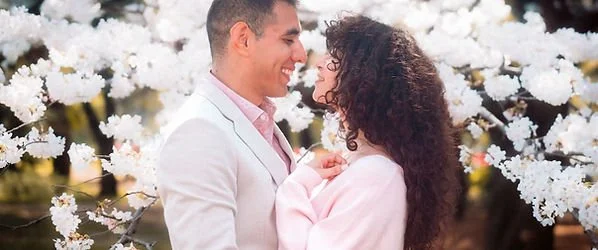 A couple smiling and looking at each other amid white flowering trees.