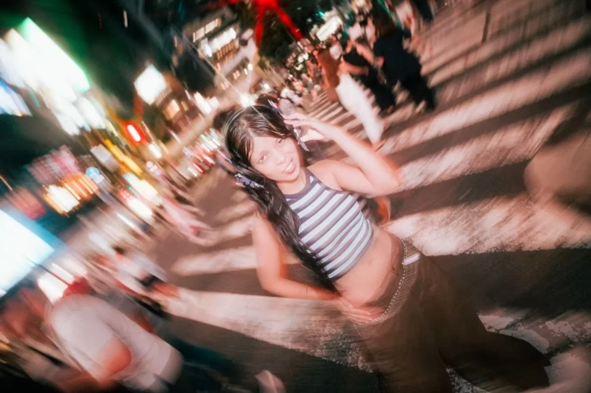 A motion-blurred image of a woman walking energetically on a busy street at night. The surrounding neon lights create a lively, bustling atmosphere as she moves, capturing the essence of city nightlife and youthful energy.