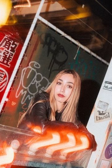 A young woman stands next to a graffiti-covered wall, holding a slice of pizza. The neon-lit surroundings enhance the vibrant street culture, with a candid and relaxed vibe that showcases a fun, urban lifestyle.