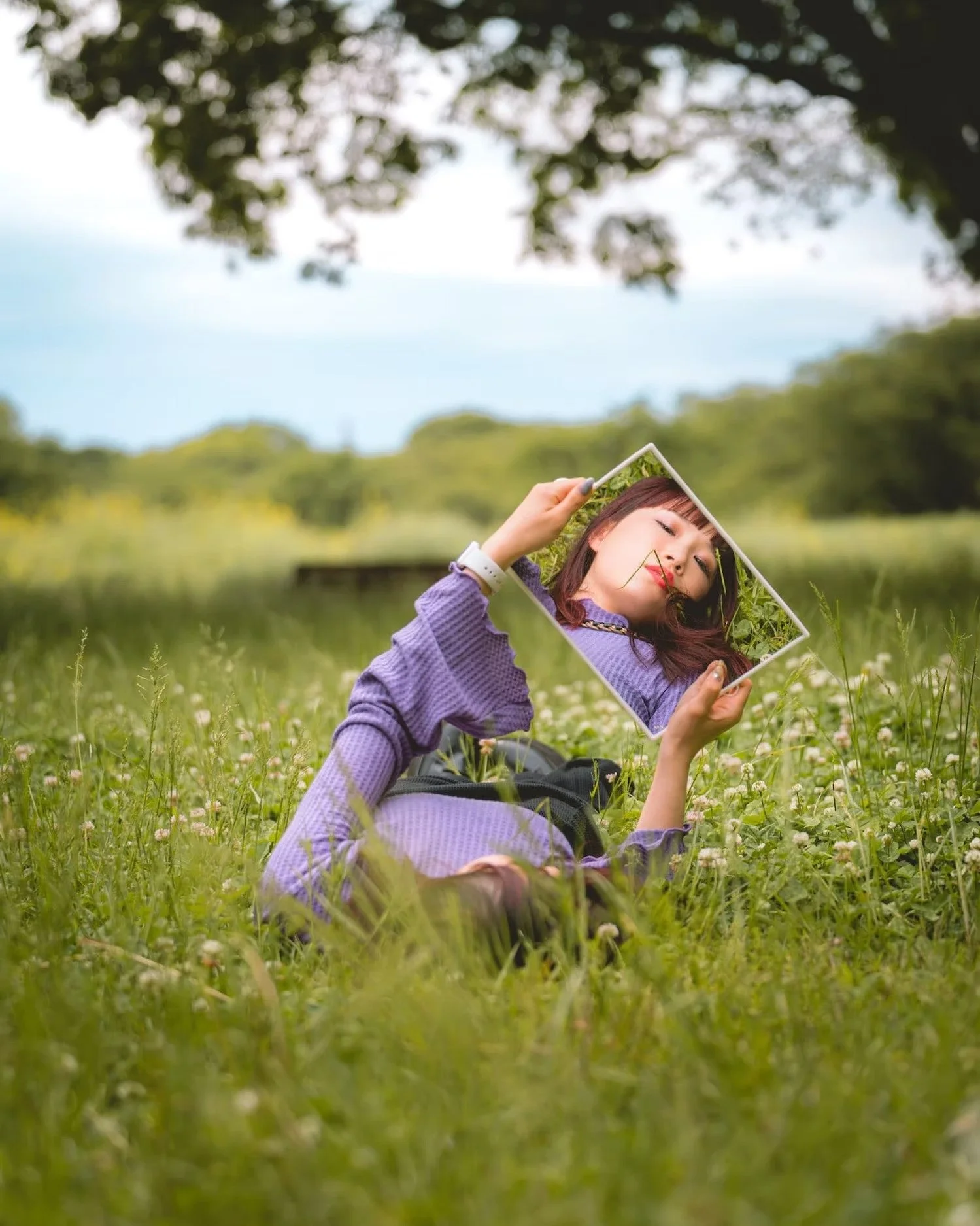 A girl sitting in a grassy field holding a mirror that reflects her face and hair, with trees and a blue sky in the background.
