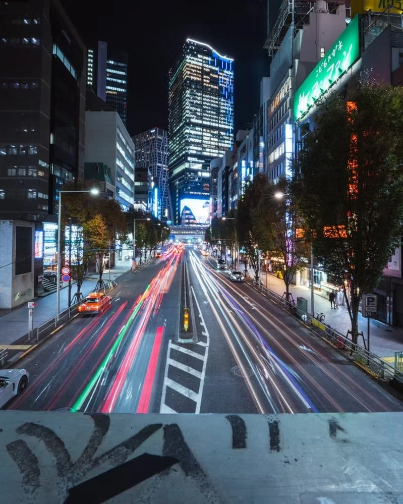 Nighttime city street scene with blurred lights of moving vehicles, tall illuminated skyscrapers, and neon signs on buildings.