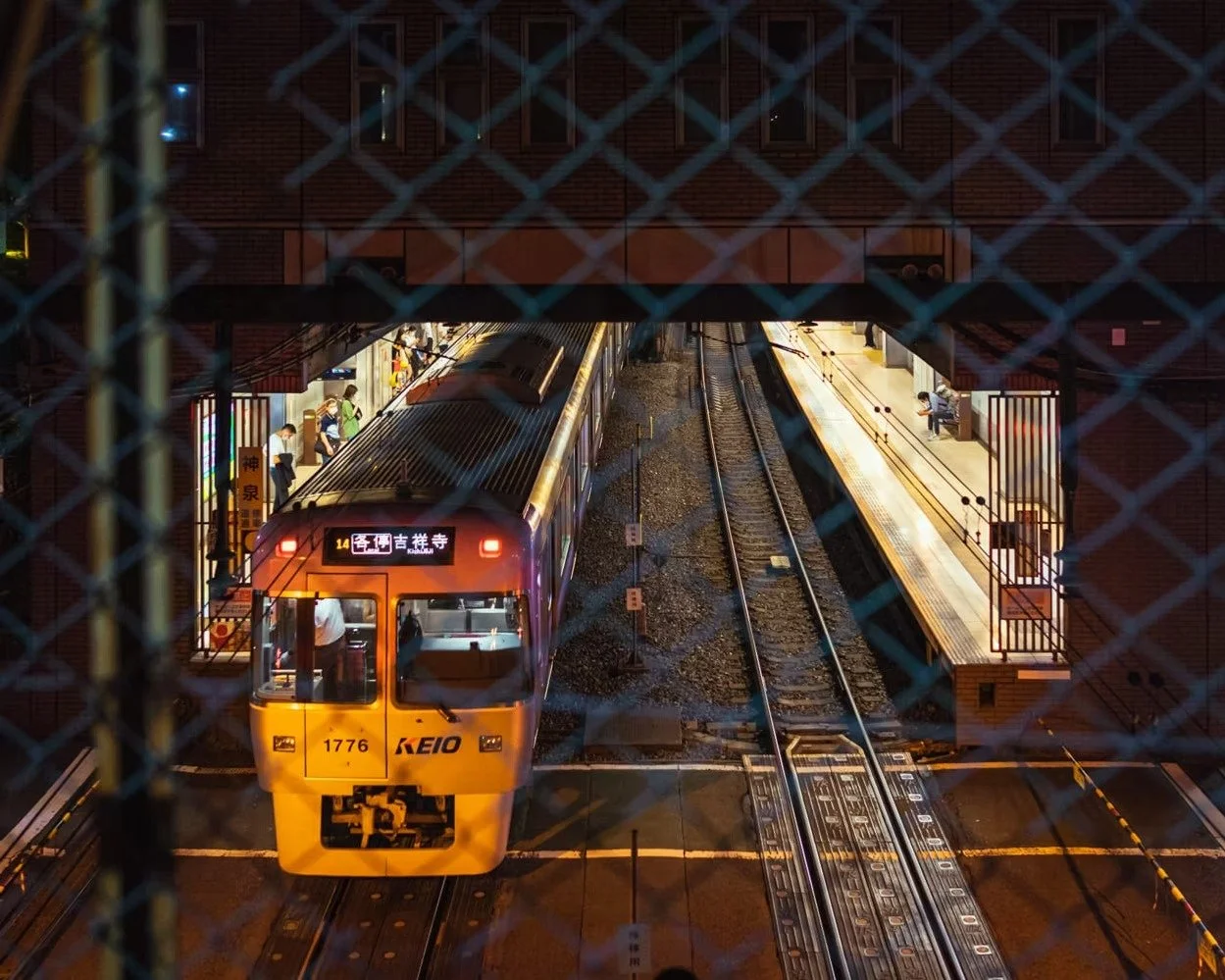 View of a train station platform at night with an arriving train viewed through a fence. Passengers are waiting on the platform.