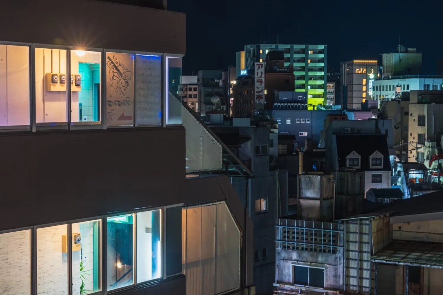 Nighttime cityscape with illuminated buildings, including a modern building with large windows and various colorful neon signs in the background.