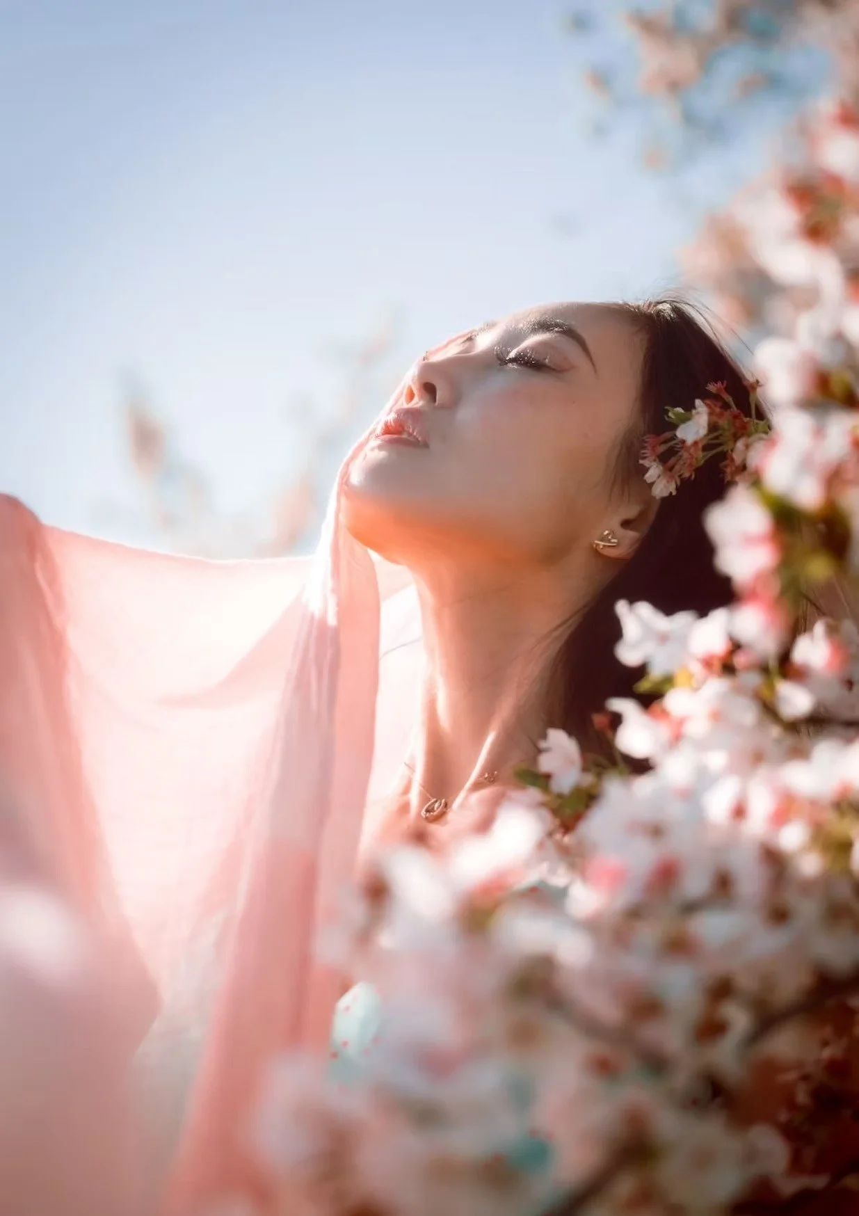 A woman stands among cherry blossoms, holding a pink scarf gently in the air. She has a peaceful expression on her face, eyes closed, as she takes in the beauty of the blooming flowers around her. The soft, dreamy light adds to the serene atmosphere.