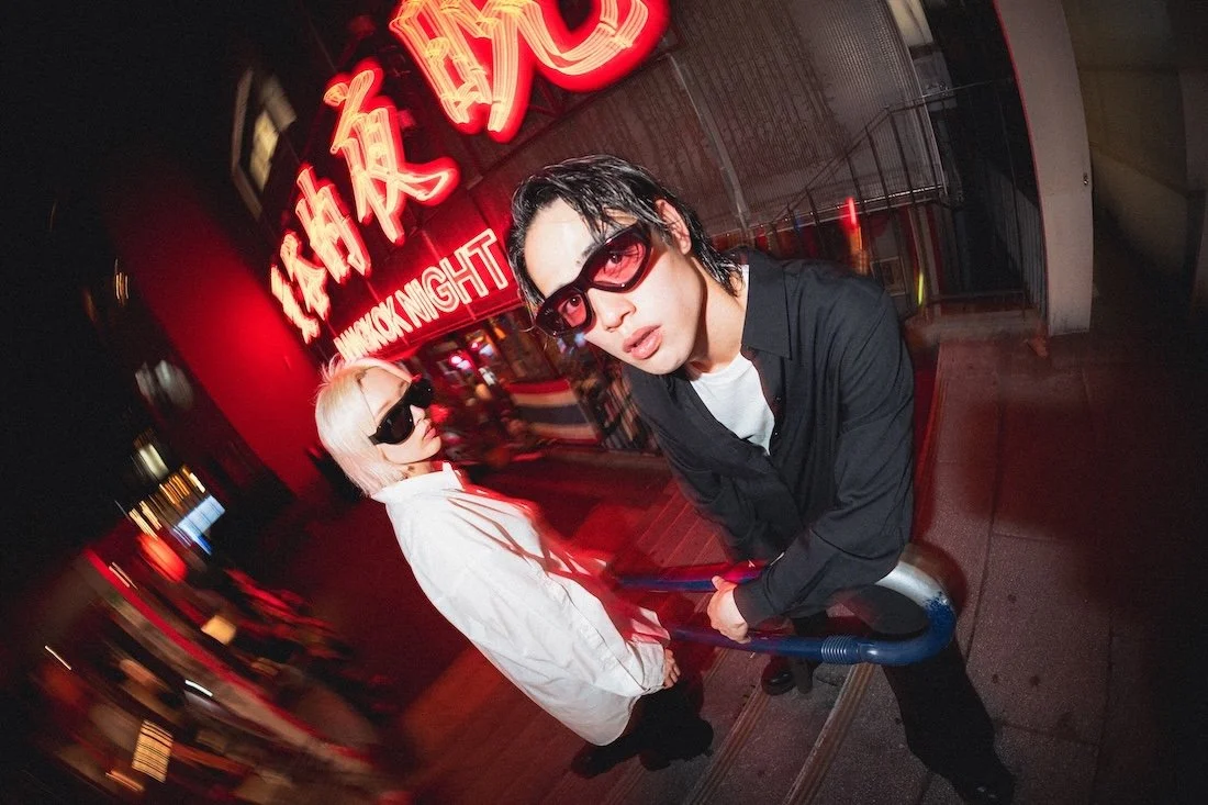 Vasuma Eyewear
---
Two young people with dark sunglasses posing at night in front of a red neon sign that reads "Midnight" in kanji characters and English, on a city street. Shot in Shibuya by fashion photographer Deniz Demir.