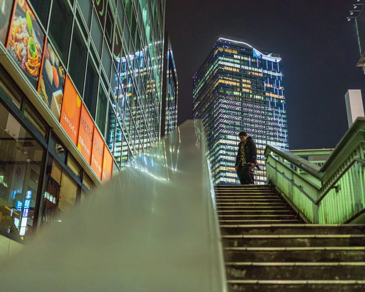 Nighttime city scene with tall modern lit-up buildings, a staircase with green railing, and a person standing on the stairs near the top.