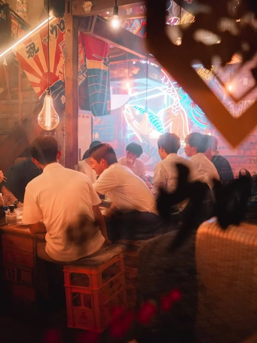 A group of young men sitting around a wooden table in a neon-lit Asian restaurant or bar, with colorful murals and fabric flags in the background, and warm lighting creating a cozy atmosphere.