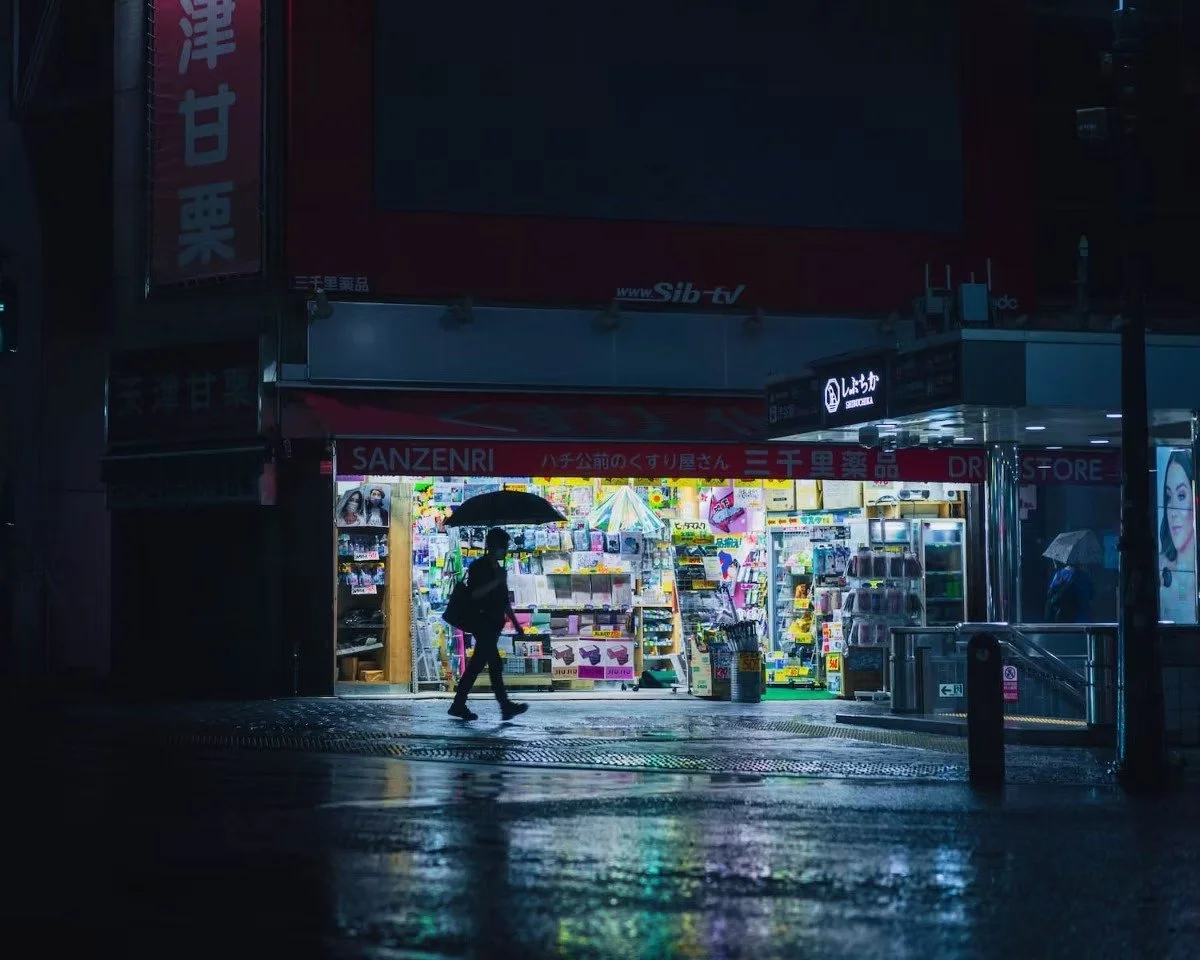 A small brightly lit drugstore in an urban area at night, with a person walking by holding an umbrella, and another person standing outside with an umbrella, reflecting a wet street in Shibuya during the Covid pandemic.