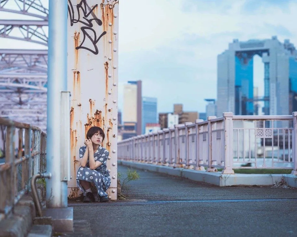 A girl sitting on a bridge in Osaka, with the city skyline in the background, creating a vibrant portrait.