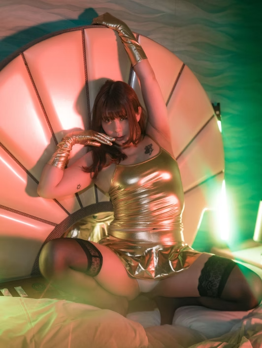 A woman wearing a shiny gold dress and matching gloves sitting on a bed with multicolored lighting.