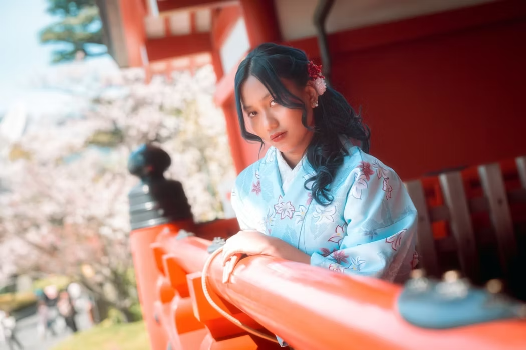 A woman wearing a traditional blue kimono with floral designs sits on a red wooden railing, with cherry blossoms in the background, at a shrine or temple in Japan.