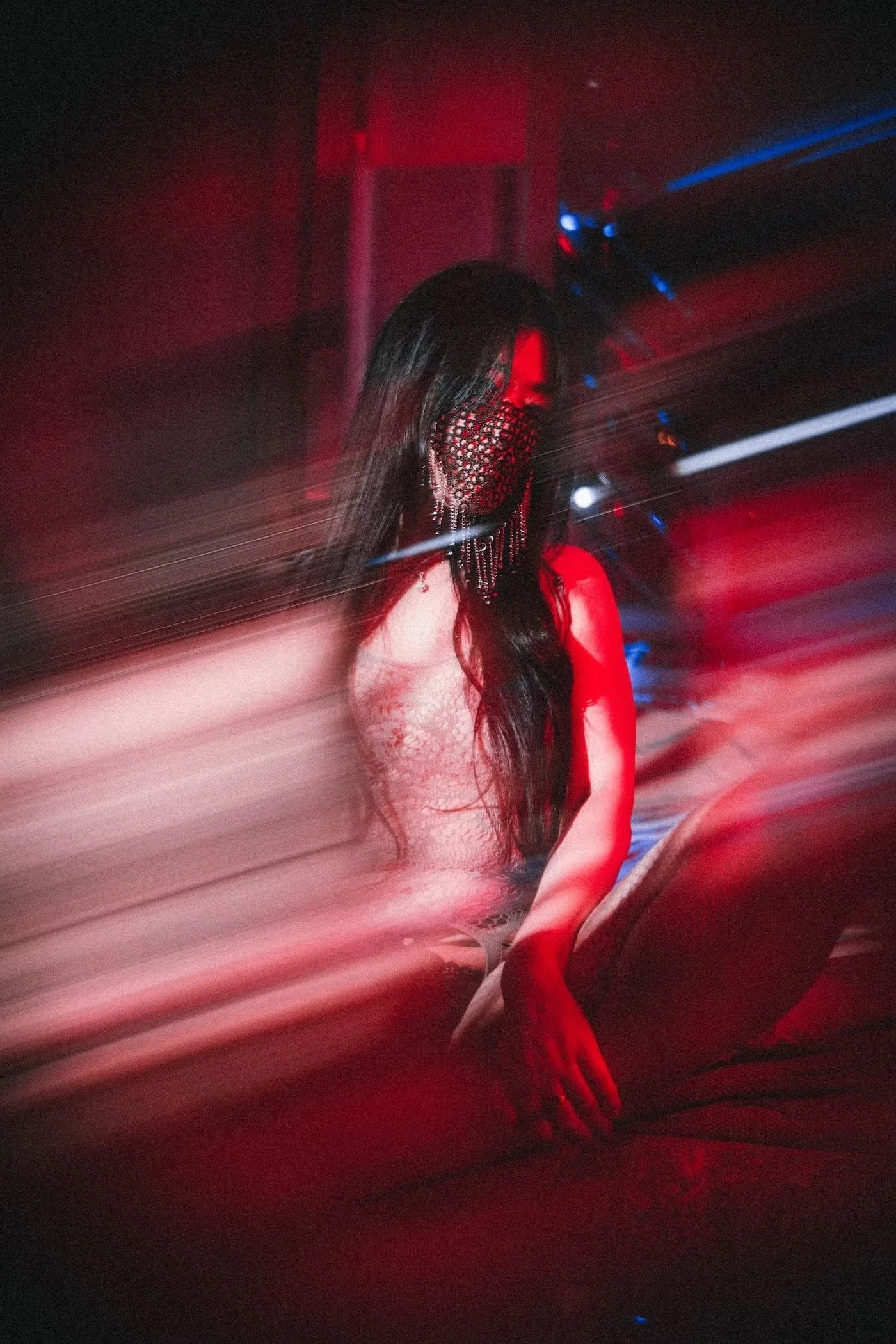 A woman with long dark hair and earrings, wearing a black fishnet face mask and lace clothing, is in a dark environment with red and blue lights creating a blurred, dynamic effect.