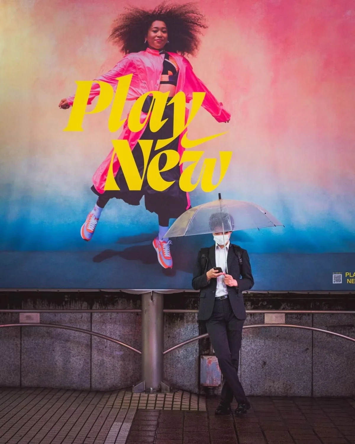 A Japanese man in a suit and face mask stands at a bus stop holding a transparent umbrella and looking at his phone. Behind him is a large colorful advertisement for Nike featuring a woman jumping in the air with the words "Play New" written in bold 