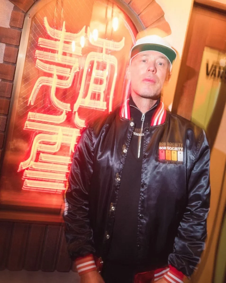 A man in a sleek varsity jacket and cap stands in front of a glowing red neon sign, exuding confidence and street style. The vibrant lights and urban backdrop create a bold, edgy atmosphere. Street photography by Deniz Demir.