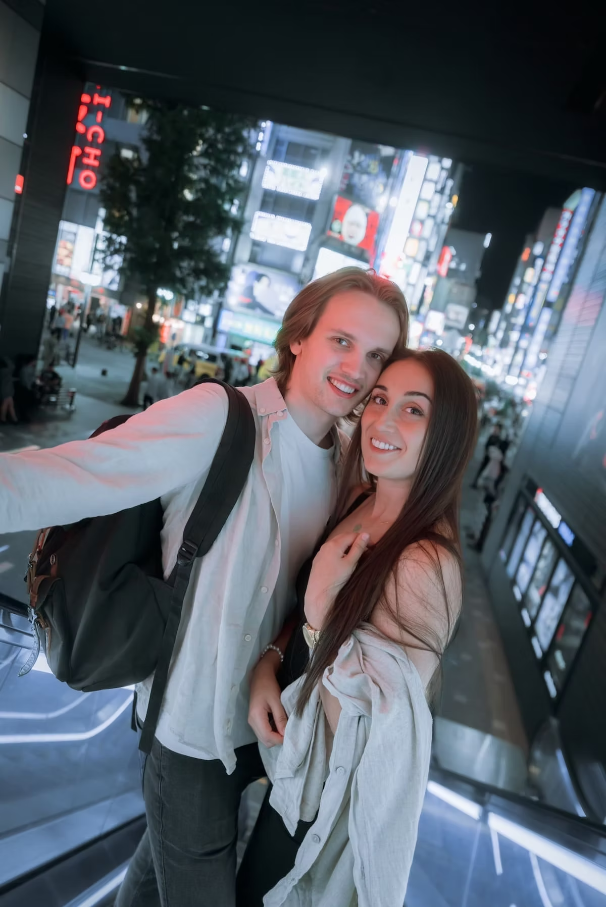 Couple smile and pose for a photo in the bustling streets of Tokyo at night. They stand close to each other, surrounded by neon lights and advertisements, capturing the energetic city vibe.