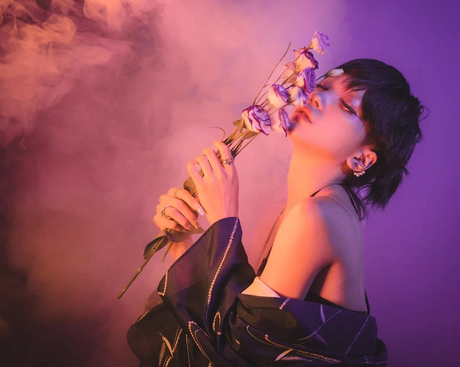 A woman with short black hair and earrings holding a bouquet of purple and white flowers with pink and purple lighting effects.