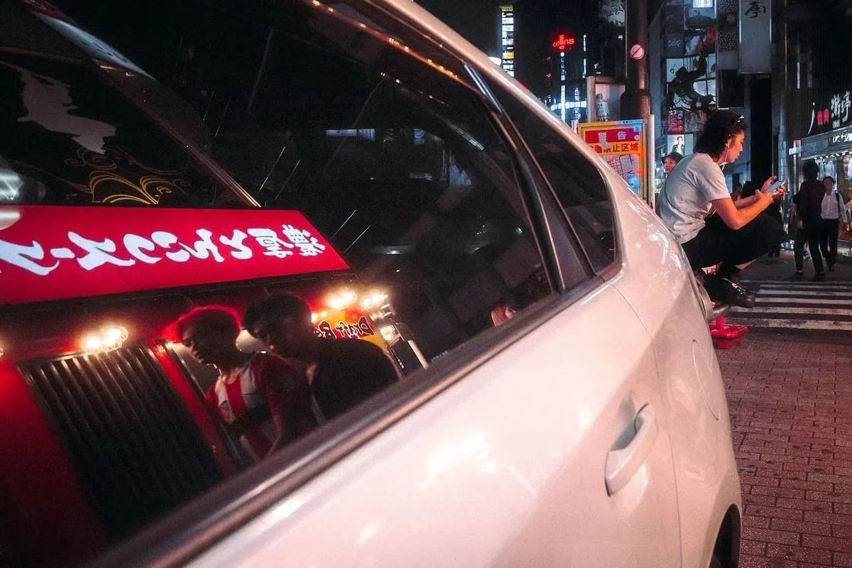 A street scene at night with a white car parked on the sidewalk, a woman sitting on the curb looking at her phone, and pedestrians walking in the background with illuminated signs. Kabukicho Shinjuku by Tokyo street photographer Deniz Demir.
