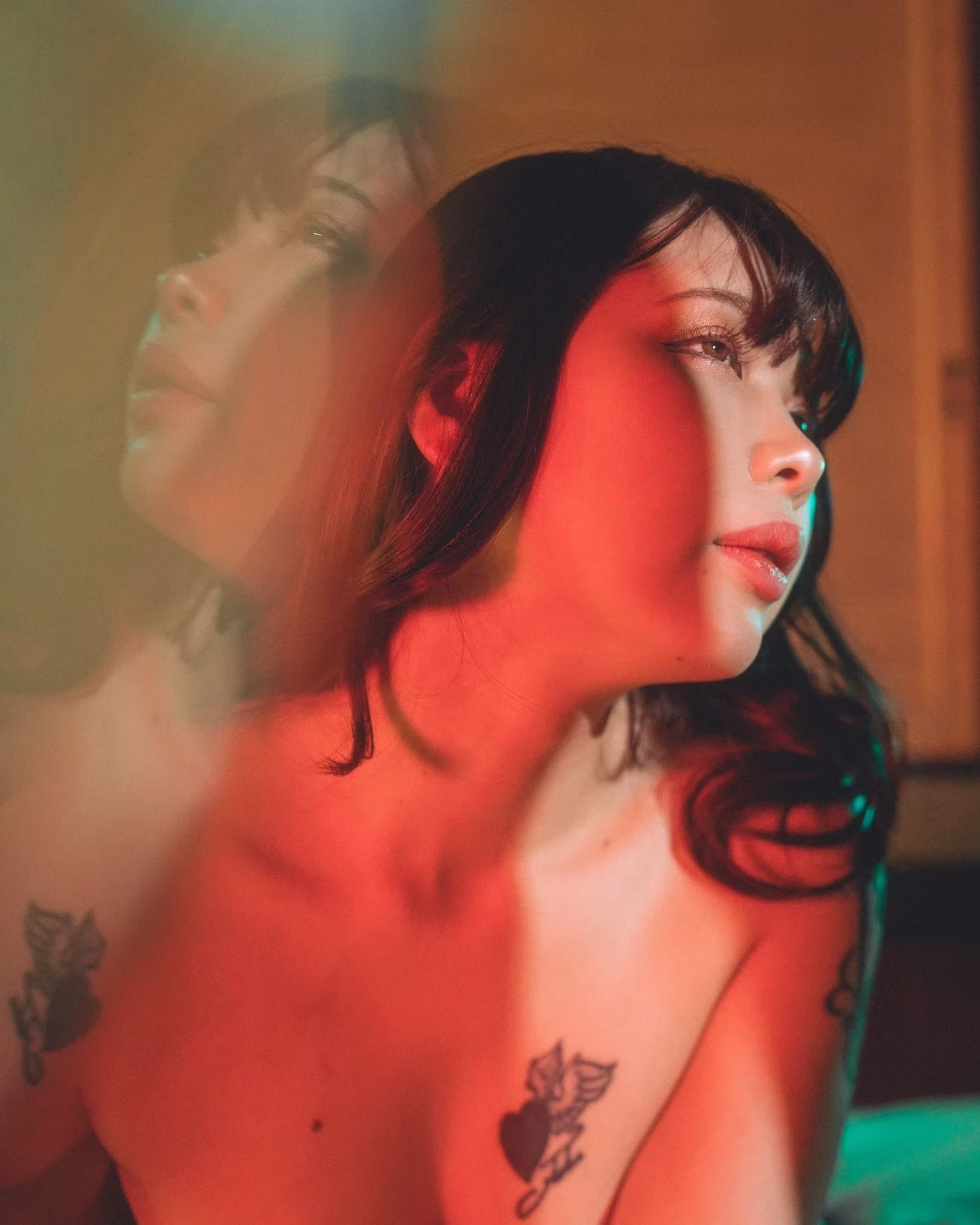 A woman with dark hair and tattoos on her shoulder and chest, looking to the side, with red and green lighting cast on her skin.