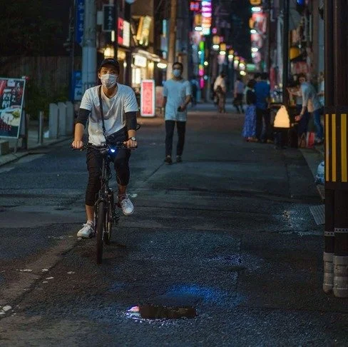 A masked cyclist rides through a neon-lit Tokyo alley at night, dodging a small puddle reflecting blue lights, with pedestrians in the background.